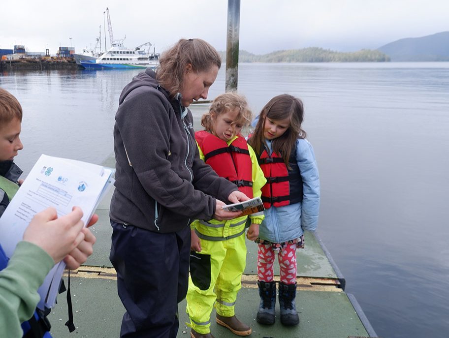 Youths wearing life vests stand on a dock as they fill out forms while an adult explains the process